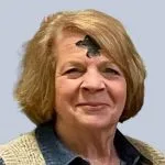 Smiling woman with short light brown hair wearing a denim shirt and patterned vest, with a cross of ashes marked on her forehead for Ash Wednesday, standing against a plain light background.