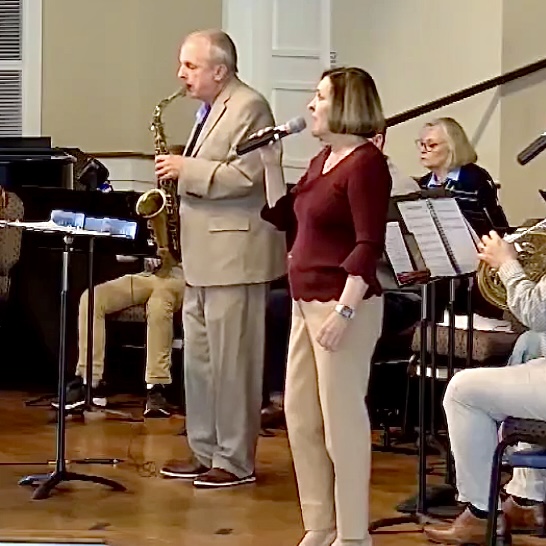 Musicians performing indoors with a small ensemble; a woman sings into a microphone while a man beside her plays the saxophone, accompanied by others on piano, horn, and woodwinds.