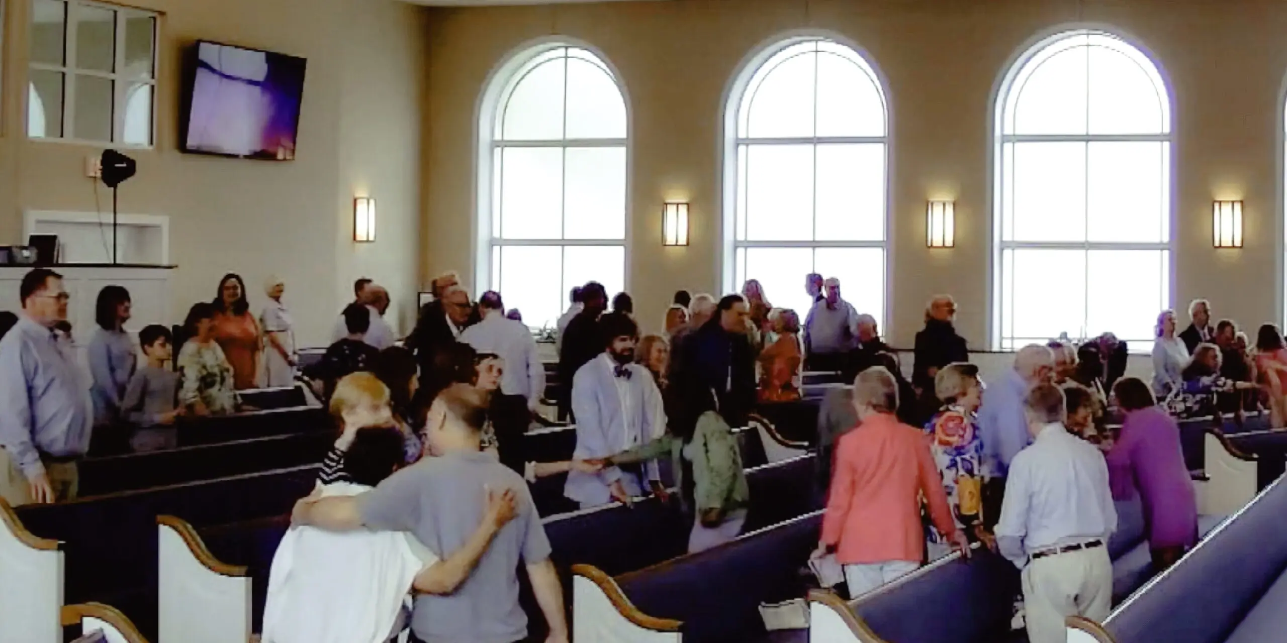 Congregation greeting one another inside a church sanctuary with tall arched windows in the background.