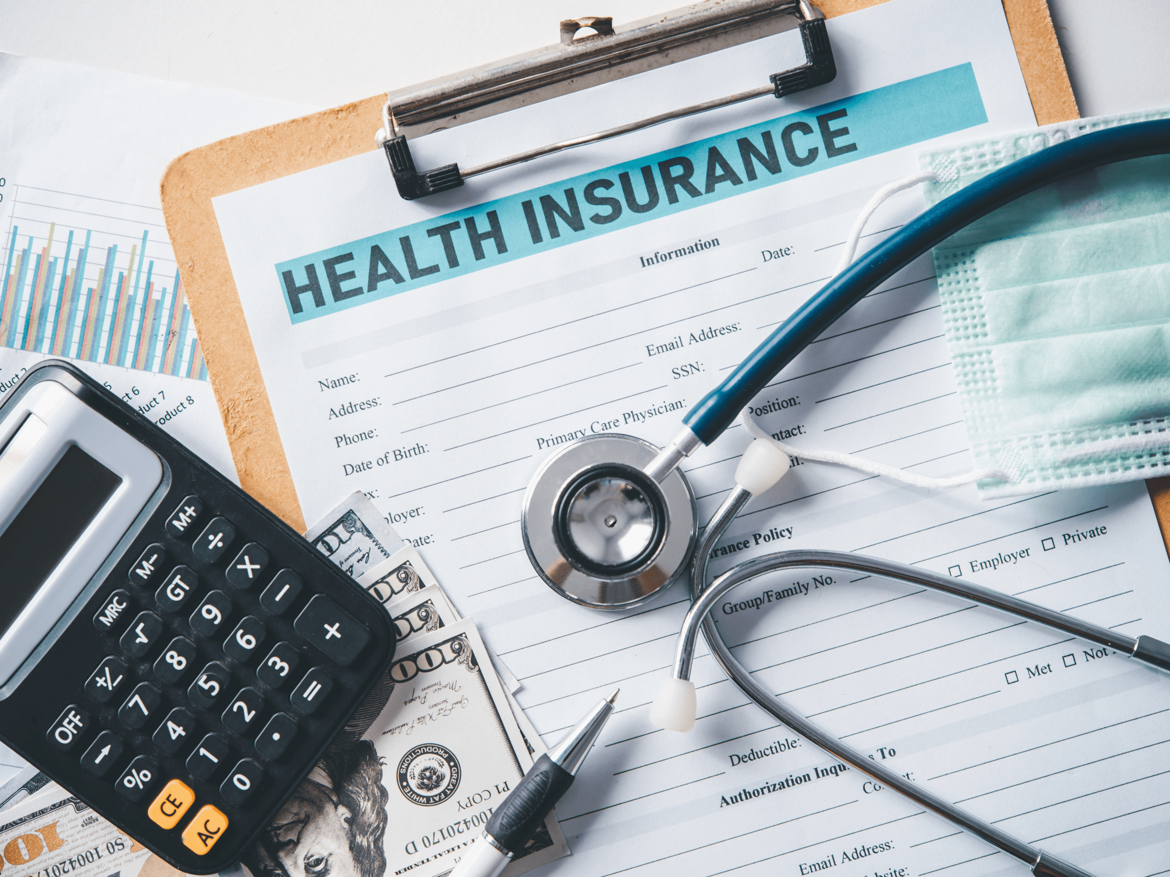 A health insurance form on a clipboard surrounded by a stethoscope, protective face mask, calculator, pen, charts, and scattered U.S. hundred-dollar bills, representing the costs and administrative details of healthcare coverage.